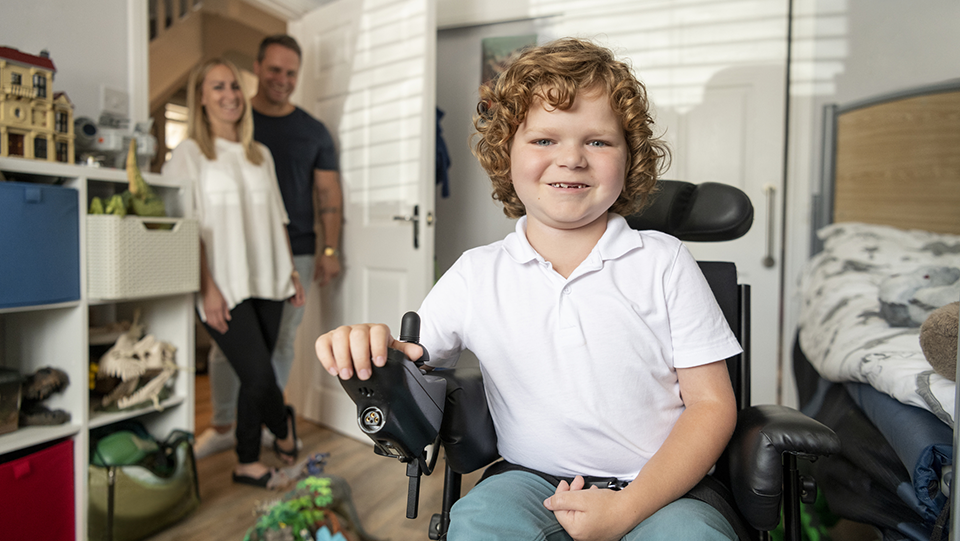 En glad pojket sitter i sin rullstol, föräldrar finns i bakgrunden